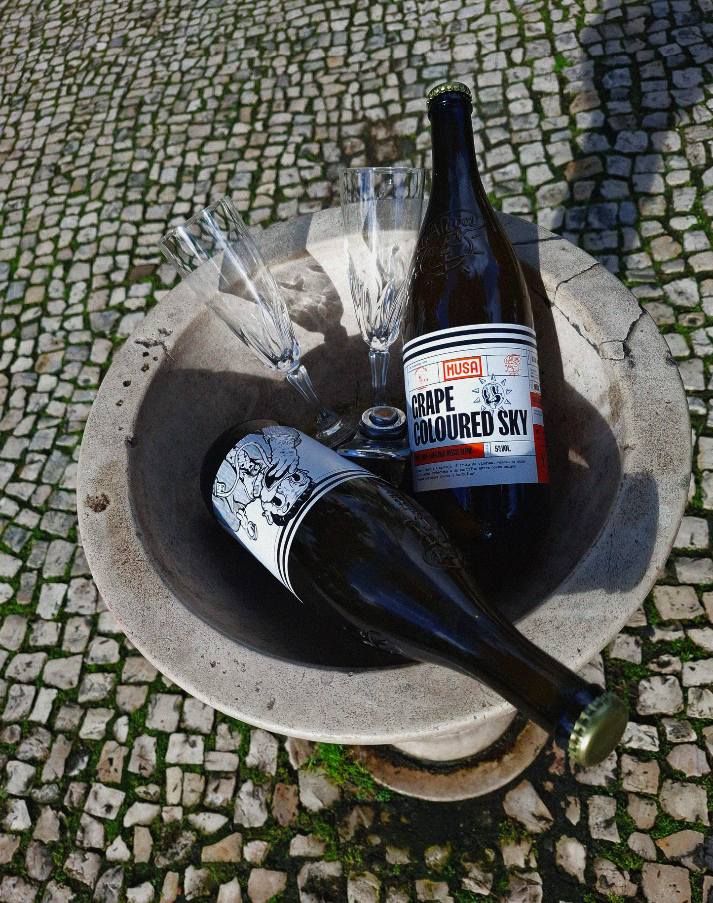 Imagem de uma garrafa de Grape Colored Sky - Grape Ale, colaboração Cerveja MUSA, Fermentage e Nat’Cool, a ser segurada numa mão com uma flute de champanhe, e o céu atrás