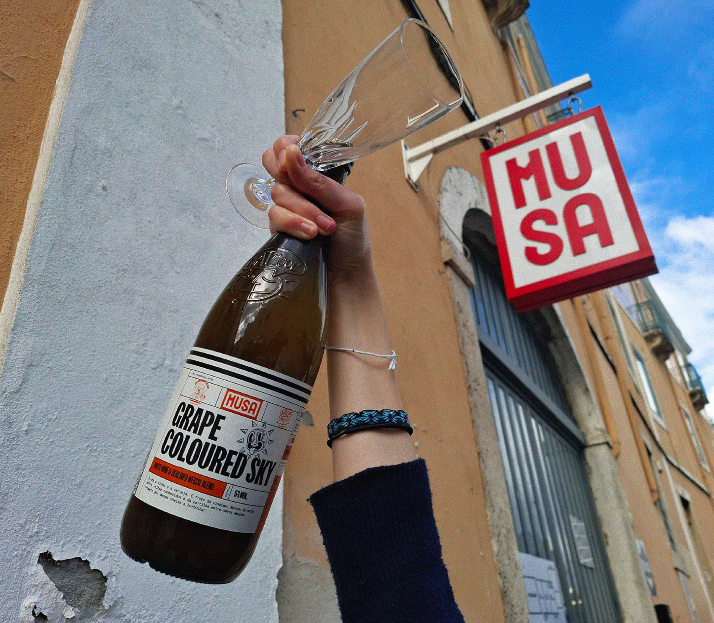 Imagem de uma garrafa de Grape Colored Sky - Grape Ale, colaboração Cerveja MUSA, Fermentage e Nat’Cool, a ser segurada numa mão com uma flute de champanhe, e o céu atrás