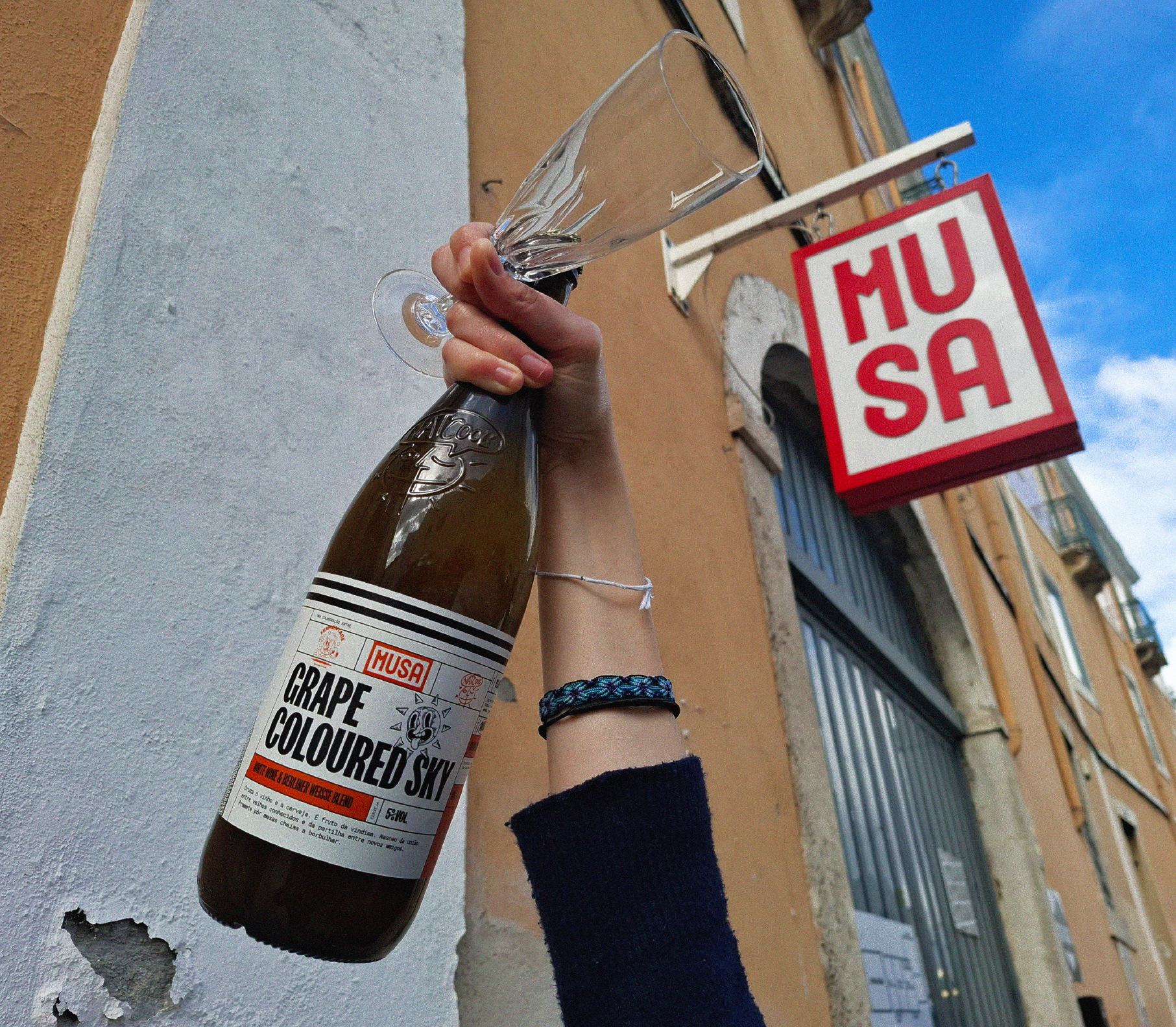 Imagem de uma garrafa de Grape Colored Sky - Grape Ale, colaboração Cerveja MUSA, Fermentage e Nat’Cool, a ser segurada numa mão com uma flute de champanhe, e o céu atrás