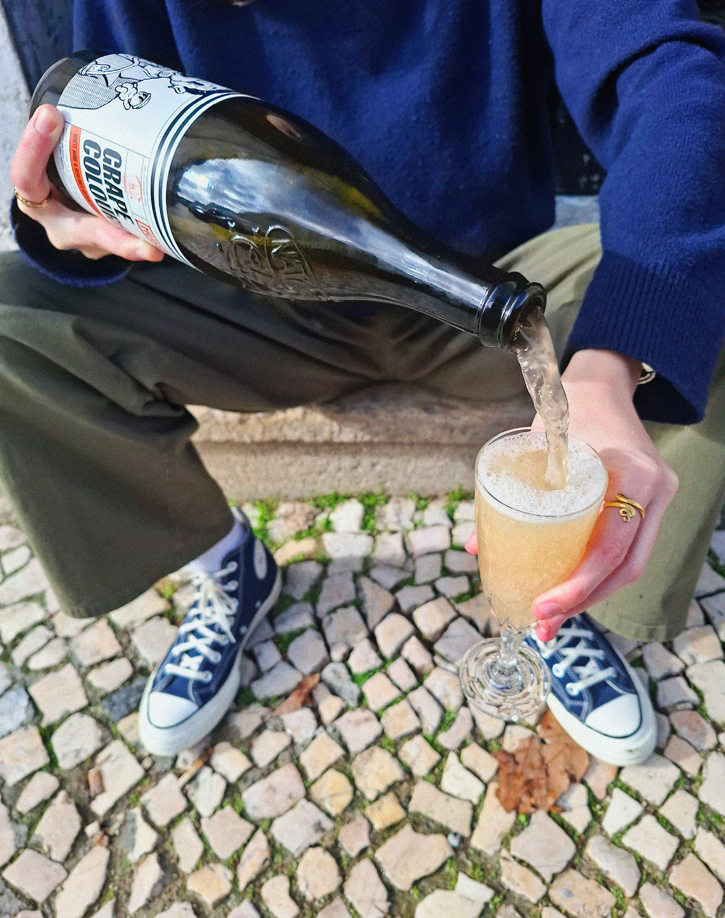 Imagem de uma garrafa de Grape Colored Sky - Grape Ale, colaboração Cerveja MUSA, Fermentage e Nat’Cool, a ser segurada numa mão com uma flute de champanhe, e o céu atrás