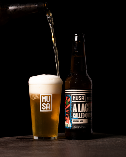 Imagem de uma garrafa de MUSA “A Lager Called Quest” (Mexican Lager) ao lado de
um copo com a cerveja, com uma espuma fofa no topo. O fundo preto destaca o rótulo
vibrante da garrafa, que apresenta uma arte colorida, e o copo está marcado com o
logótipo da MUSA